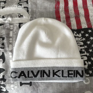 Vit mössa från Calvin Klein - Vit mössa från Calvin Klein med svart logotyp och randig detalj på uppviket. Snygg och stilren design som passar perfekt till kalla dagar. Tillverkad i mjukt material för skön komfort. Perfekt accessoar för dig som gillar klassisk streetstyle. Pris kan diskuteras
