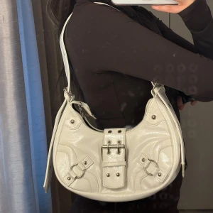 Vit axelväska med spänndetaljer - Säljer en snygg vit axelväska i glansigt skinnliknande material med coola silverfärgade spännen och ringar framtill. Väskan har en bågformad design och justerbar axelrem. Perfekt för att lyfta vilken outfit som helst.