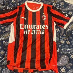 AC Milan Puma tröja  - AC Milan fotbollströja från Puma med klassiska röda och svarta vertikala ränder, vita detaljer och klubbmärke på bröstet. Tryckta sponsorer på framsidan och ärmen. Tillverkad i lätt och ventilerande polyester, perfekt för match eller träning.