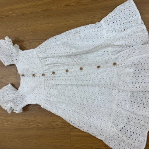 Vit broderad sommarklänning med volang - Superfin vit klänning i bomull med broderade hål och volangdetaljer. Klänningen har korta puffärmar, knappar framtill och en luftig, flowy passform. Perfekt för varma dagar och ger en romantisk vibe. (Barn klänningar) 