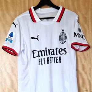 AC Milan vit bortatröja Puma Theo 19 - AC Milan bortatröja i vitt med svarta och röda detaljer, tillverkad av Puma. Kortärmad modell med klubbmärke, sponsortryck och Serie A-logga. Namn och nummer Theo 19 på ryggen. Snygg krage och ärmslut med röd och svart kant. Perfekt för fans av italiensk fotboll.