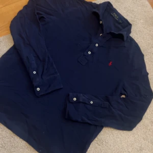 Marinblå långärmad pikétröja Polo Ralph Lauren - Snygg marinblå långärmad pikétröja från Polo Ralph Lauren med klassisk krage, knappar framtill och röd broderad logga på bröstet. Tillverkad i 100% bomull för en skön och avslappnad känsla. Perfekt till jeans eller chinos. Kommer strykas efter köp