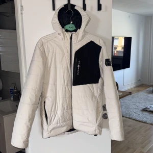 Vit och svart pufferjacka Sail Racing - 🔔Priset kan diskuteras                         Snygg pufferjacka från Sail Racing i vit med svarta detaljer. Jackan har huva, dragkedja och flera fickor. Perfekt för kalla dagar med sin vadderade design och sportiga look. Stora Sail Racing-loggor på bröst och ärm. Riktigt clean och modern stil.