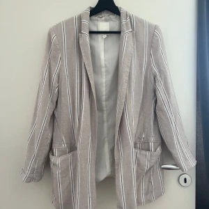 Randig beige kavaj från H&M, strl 44 - Snygg randig kavaj från H&M i beige med vita och svarta ränder. Oversized känsla, två fickor framtill och klassisk krage. Tillverkad i en blandning av linne och viskos. 