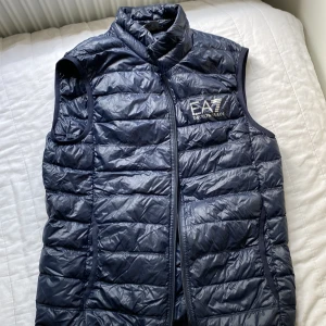 Mörkblå dunväst från EA7 Emporio Armani - Snygg mörkblå dunväst från EA7 Emporio Armani med quiltad design och dragkedja framtill. Västen har hög krage och logga broderad på bröstet. Perfekt att slänga över en hoodie eller tröja för extra värme och stil.