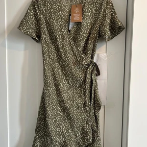 Grön omlottklänning från Vero Moda - Supersöt grön omlottklänning från Vero Moda med vitt leopardmönster. Klänningen har korta ärmar, volangkant nertill och knyt i midjan. Tillverkad i 100% återvunnen polyester – perfekt för dig som gillar både stil och hållbarhet.