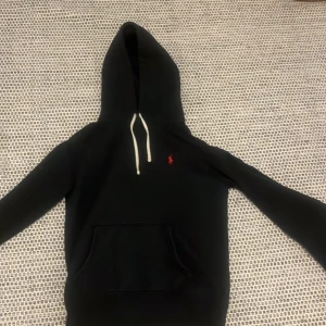 Svart hoodie från Polo Ralph Lauren - Svart hoodie från Polo Ralph Lauren med klassisk röd broderad logga på bröstet. Tröjan har huva med vita snören och en stor magficka. Perfekt för dig som gillar stilren och enkel design med en touch av lyx.