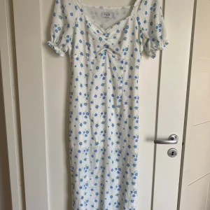 Vit blommig långklänning NA-KD - Supersöt långklänning från NA-KD x Queen of Jetlags i vitt med blått blommönster. Klänningen har korta puffärmar med volangkant, v-ringning och lätt rynk framtill. Perfekt för sommarens alla tillfällen!