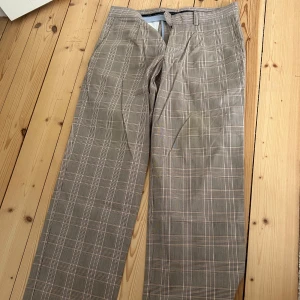Rutig kostymbyxa Tommy Hilfiger 32 - Snygga rutiga kostymbyxor från Tommy Hilfiger Golf i beige och bruna toner med inslag av orange. Klassisk rak passform med bälteshällor och diskret dragkedja. Perfekta för en stilren och trendig look.