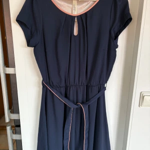 Mörkblå klänning från Broadway NYC - Mörkblå kortklänning med rosa kantdetaljer från Broadway NYC. Klänningen har korta ärmar, nyckelhålsöppning vid halsen och ett smalt bälte i midjan. Tillverkad i ett lätt och mjukt material, perfekt för en stilren look.