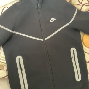 Svart Nike kofta med reflexdetaljer - Svart hoodie från Nike med gråa reflexdetaljer på bröst, rygg och fickor. Tröjan har dragkedja framtill, två fickor med dragkedja och huva. Perfekt för dig som vill sticka ut med snygg sportig stil och synas i mörkret.