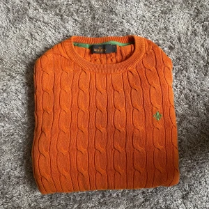 Orange kabelstickad tröja Morris M - Snygg orange kabelstickad tröja från Morris i 100% merceriserad bomull. Tröjan har rund halsringning och en grön broderad logga på bröstet. Perfekt för dig som vill sticka ut med färg och stil i höst eller vår.
