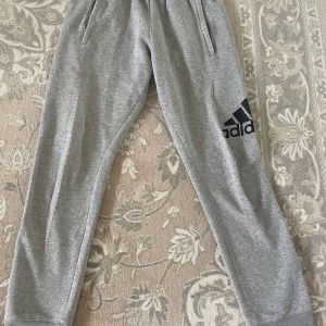 Grå mjukisbyxor från Adidas S - Grå mjukisbyxor från Adidas med svart logga på benet. Byxorna har resår i midjan, sidofickor och en bakficka. Tillverkade i mjukt bomullsmaterial, perfekta för chill eller träning. Klassisk sportig look och bekväm passform.den en rosa fläkt som ni ser på sista bild 