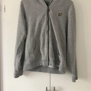 Grå hoodie från Lyle & Scott - Grå hoodie från Lyle & Scott med dragkedja och klassisk gul logga på bröstet. Tröjan har huva, känguruficka och ribbade muddar. Perfekt för chill dagar eller när du vill ha en enkel och stilren look.