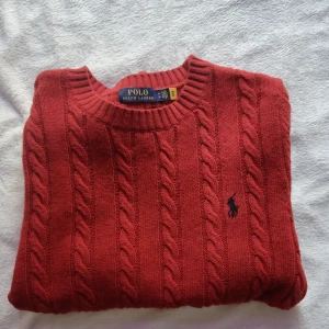 Röd kabelstickad tröja Polo Ralph Lauren - Kabelstickad röd tröja från Polo Ralph Lauren med klassisk rund halsringning och broderad logga på bröstet. Tillverkad i en mix av ull, akryl och nylon för både värme och komfort. Perfekt för dig som gillar en tidlös och stilren look.