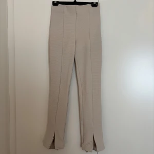 Beige ribbade byxor med slits - Snygga beige byxor med ribbad struktur och raka ben. Byxorna har hög midja och slits framtill vid bensluten för en trendig look. Perfekta att styla med sneakers eller boots. Materialet känns mjukt och stretchigt. Finns en fläck längst ner på slitsen!