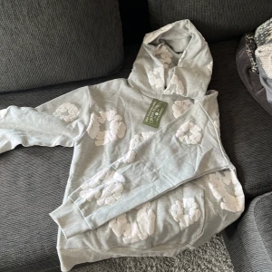 Denim tears hoodie - Säljer en grå hoodie från Denim Tears med unika vita molnliknande tryck över hela tröjan. Tröjan har huva och långa ärmar, perfekt för dig som gillar streetwear och vill sticka ut med en cool design. Materialet känns mjukt och skönt mot huden.