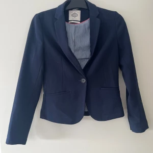 Marinblå kavaj ifrån bershka - Marinblå kavaj ifrån bershka i storlek M. Den är figursydd, har en knapp framtill och två fickor utan lock. Ärmarna har två knappar vid ärmsluten, som man kan förstora med. Kavajen är kort i modellen och slutar ungefär vid midjan. En klassisk och stilren modell som passar både till vardags och festligare tillfällen. Kontakta för övriga frågor.