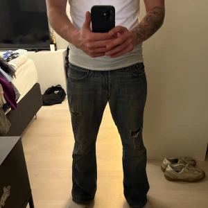 Levi's 527 bootcut jeans blå - Levi's 527 low bootcut jeans i klassisk blå tvätt. Jeansen har slitna detaljer och hål på benen samt bakfickan för en rå, vintage vibe. Tillverkade i 100% bomull med bootcut passform och låg midja. Perfekta för dig som gillar en avslappnad stil. 