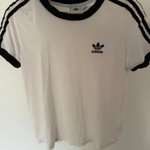 Vit Adidas t-shirt med svarta detaljer - Vit t-shirt från Adidas med svarta ribbade muddar vid hals och ärmslut samt svarta ränder längs axlarna. Klassisk Adidas-logga broderad på bröstet. Skön passform och sportig stil.