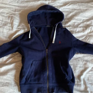 Mörkblå hoodie från Polo Ralph Lauren - Säljer en mörkblå hoodie från Polo Ralph Lauren med dragkedja och huva. Tröjan har vita snören vid huvan, två fickor framtill och en liten röd logga broderad på bröstet. Perfekt för en avslappnad stil. Finns tecken på användning, finns fläckar vet ej om de går bort i tvätten och ett litet hål på baksidan av armen men det syns knappt.
