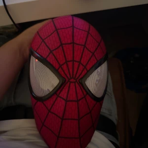 Spiderman mask med avtagbara ögon - Cool Spiderman-mask i röd och svart med klassiskt nätmönster och stora vita avtagbara ögon. Masken har dragkedja baktill och är tillverkad i stretchigt syntetmaterial för en tight passform. Perfekt för cosplay eller maskerad!