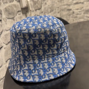 Blå bucket hat från Dior - Snygg bucket hat från Dior i blått och vitt med ikoniskt Dior-mönster över hela hatten. Tillverkad i 100% bomull och fodrad med blått tyg på insidan. Perfekt accessoar för att lyfta din stil med en lyxig touch.