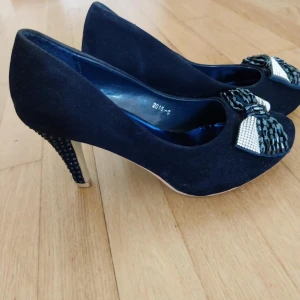 Svarta pumps med rosett - Stilrena svarta pumps i mocka med hög klack och snygg rosettdetalj framtill i svart och silver. Klacken är dekorerad med små svarta pärlor för extra bling. Perfekta för dig som vill sticka ut med elegans.