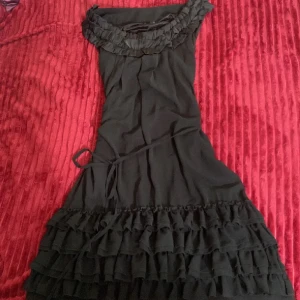 Svart volangklänning med knytband - Supersöt svart kortklänning med flera lager volanger nertill och volangdetaljer vid halsringningen. Klänningen har ett smalt knytband i midjan och är tillverkad i ett lätt, flowy tyg med satinfoder. Perfekt för dig som gillar romantiska vibes och vill sticka ut.