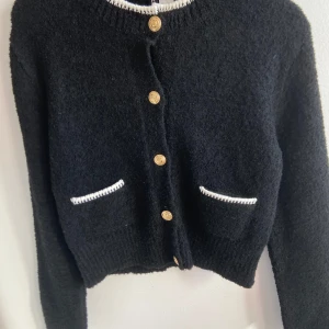 Festlig stickad svart kofta - Stilren stickad svart kofta med guldiga knappar framtill och två fickor med vit kant. Pris kan diskuteras 