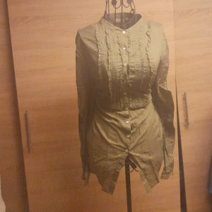 Olivgrön blus med volang från MyWear - Olivgrön blus från MyWear med långa ärmar och snygga volangdetaljer framtill. Skjortan har knappar hela vägen och en feminin passform. Perfekt för dig som gillar en stilren look med lite extra detaljer.