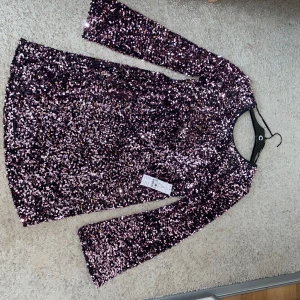 Glittrig lila paljettklänning från Gina - Superglittrig lila kortklänning från Gina med långa ärmar och täckt av paljetter. Perfekt för dig som vill sticka ut och glänsa. Klänningen har en rak passform och rund halsringning. Snygg till sneakers eller klackar!