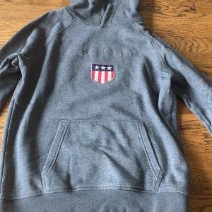 Grå hoodie från Gant med brodyr - Snygg grå hoodie från Gant med broderad logga och amerikanskt inspirerat emblem på bröstet. Klassisk känguruficka framtill och huva. Perfekt för dig som gillar stilren streetwear med sportig vibe.