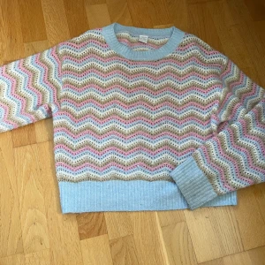 Pastellfärgad stickad tröja Lindex - Superfin stickad tröja från Lindex i pastelliga färger med vågigt mönster i ljusblått, beige och rosa. Tröjan har ribbade muddar och rund hals, perfekt för dig som gillar färg och mysiga vibes.