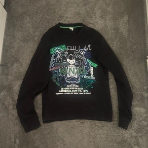 Svart Kenzo sweatshirt med tigertryck - Svart sweatshirt från Kenzo med ikoniskt broderat tigerhuvud och gröna, blå och vita detaljer framtill. Tröjan har rund hals, långa ärmar och ribbade muddar. Perfekt statement piece för dig som gillar streetstyle och coola prints.