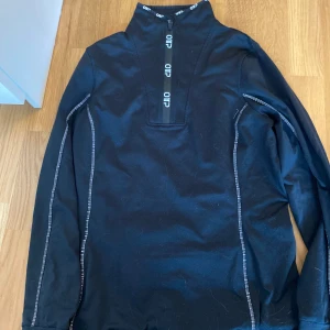 Svart långärmad  half zip tröja i strl M - Svart långärmad tröja med vit kontrastsöm längs ärmarna och halv dragkedja framtill. Hög krage med logga på dragkedjan ger en sportig vibe. Perfekt för lager-på-lager under kyliga dagar. I strl M. Kan mötas upp i Trollhättan eller Alingsås. Kan skickas men då tillkommer en fraktkostnad.