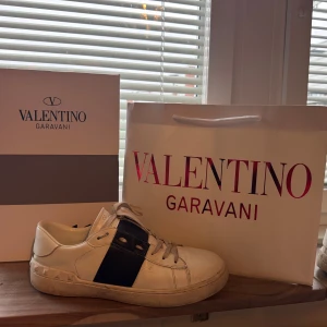 Valentino Garavani vita sneakers - Snygga vita sneakers från Valentino Garavani med bred svart rem över sidan och coola nitar på hälen. Skorna har rund tå, snörning och platt sula. Tillverkade i skinn och levereras med originalkartong och dustbag.