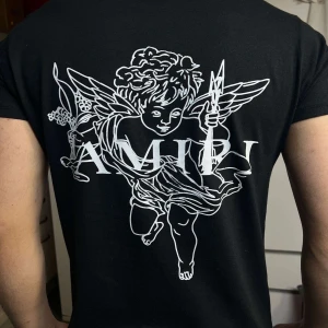 Svart AMIRI t-shirt med ängeltryck - Svart t-shirt från AMIRI i mjuk bomull med korta ärmar. Framsidan har AMIRI-logga med färgstänk, baksidan har stort vitt tryck av en ängel och loggan. Snygg och edgy design som sticker ut och passar perfekt till streetwear-stilen.