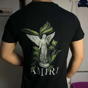 Svart Amiri t-shirt med ängeltryck - Svart t-shirt från Amiri med stor grafisk ängel och gröna blad på ryggen samt Amiri-logga. Framsidan har mindre tryck med samma motiv och logga. Klassisk passform och rund hals. Perfekt för dig som gillar streetwear med konstnärlig vibe.