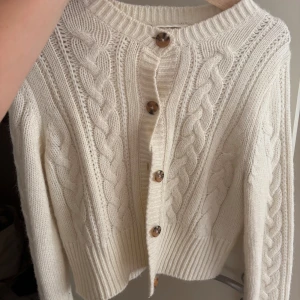 Vit kabelstickad kofta från Monki - Supermysig vit kabelstickad kofta från Monki med snygga bruna knappar framtill. Koftan har rund halsringning, ribbade muddar och lång ärm. Perfekt för lager-på-lager och chill höst- och vinterstil.