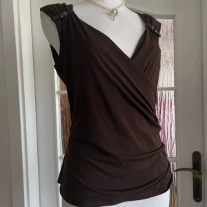Brun omlottopp från Michael Kors - Snygg brun omlottopp från Michael Kors med coola spänndetaljer på axlarna. Toppen är ärmlös och har en smickrande v-ringning och draperad front. Perfekt för dig som gillar stilrena och trendiga plagg.
