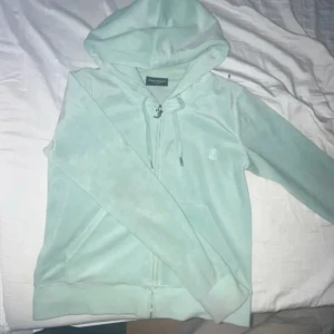 Mintgrön Juicy Couture kofta i velour - Supermjuk mintgrön kofta  från Juicy Couture i klassisk velour. Tröjan har dragkedja med snygg JC-detalj, huva med snörning och två fickor framtill. Perfekt för en chill och trendig look.