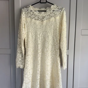 Vit spetsklänning med lång ärm - Superfin vit klänning med blommig spets och lång ärm. Klänningen har en romantisk känsla med transparenta detaljer upptill och vackert mönster över hela plagget. Perfekt för sommarens speciella tillfällen eller student.
