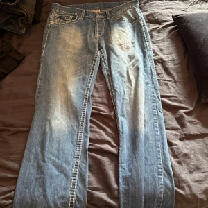 True Religion  - Säljer ett par ljusblå True Religion jeans med tydliga kontrastsömmar och ikoniska bakfickor. Klassisk femficksmodell i bomull med snyggt slitna detaljer och bred passform. Perfekta för dig som gillar streetwear och vill sticka ut.