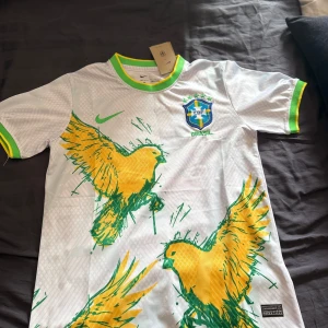 Vit Brasilien fotbollströja Nike M - Snygg vit Brasilien fotbollströja från Nike med gröna och gula detaljer. Tröjan har korta ärmar, grönt och gult ribbade muddar och krage, samt stora gula fåglar tryckta framtill. Broderat CBF-märke på bröstet och Dri-FIT-material som andas.