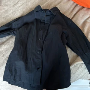 Svart klassisk skjorta i bomull - En svart skjorta med klassisk krage och knäppning framtill. Skjortan har lång ärm och är tillverkad i ett mjukt bomullstyg. Perfekt för en stilren och enkel look.