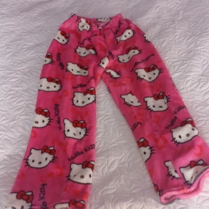 Hello Kitty fleecebyxor rosa - Supermjuka och mysiga rosa fleecebyxor med Hello Kitty-motiv över hela byxorna. Byxorna har resår i midjan och är perfekta för chill hemma. Sött tryck med röda rosetter och vita Hello Kitty-ansikten. Perfekt för dig som älskar kawaii-stil!