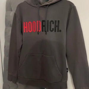 Grå HOODRICH hoodie med tryck - Säljer en grå hoodie från HOODRICH i storlek S. Tröjan har stor logga framtill i rött och svart, huva med snörning.
