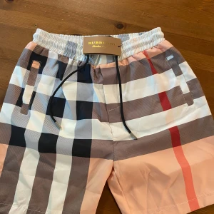 Original färg Burberry shorts - Snygga shorts från Burberry med klassiskt rutigt mönster i beige, svart, vit och röd. De har elastisk midja med svart snörning och praktiska fickor fram och bak. Perfekta för sommaren när du vill sticka ut med en lyxig och trendig look. Helt ny inte andvänd!