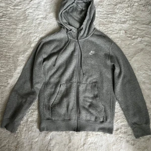 Grå Nike hoodie med dragkedja XS - Klassisk grå hoodie från Nike i storlek XS. Tröjan har huva med snörning, dragkedja framtill och två stora fickor. Tillverkad i mjuk bomullsblandning och perfekt för chill eller träning. Diskret Nike-logga på bröstet.